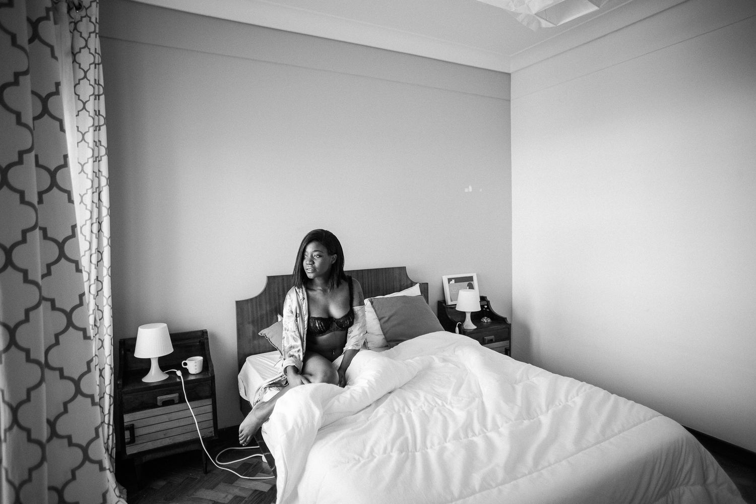 A black and white photograph of someone sitting on a bed in a hotel room with modern decor and bedside lamps.