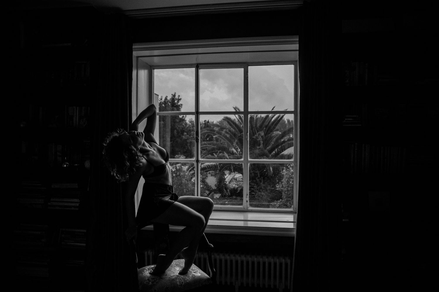 A silhouetted figure sits by a window in a dark room, with natural light streaming through the glass panes.