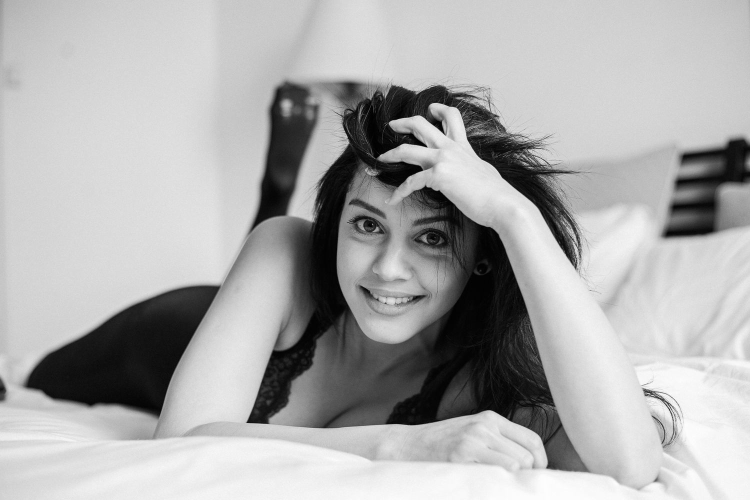 Black and white photo of someone lying on white bedding while running fingers through their dark hair.