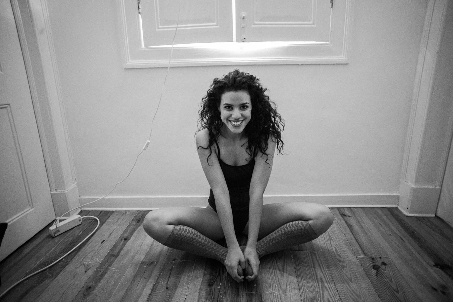 A person in dark clothing sits on a hardwood floor against a white wall in an artistic pose.