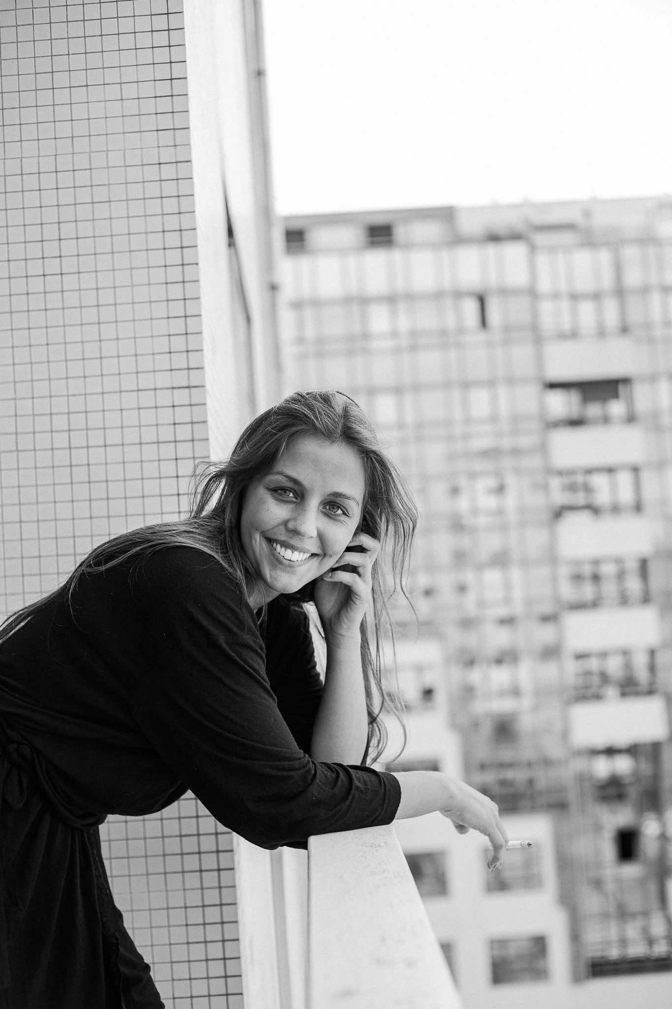A person in black clothing leans against a white tiled wall in an urban setting with buildings visible in background.