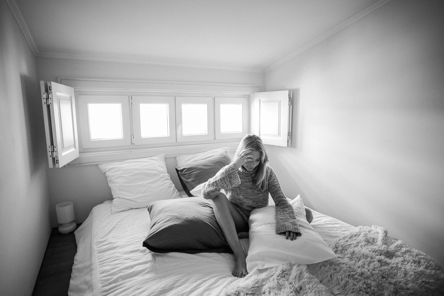 Black and white photo of someone lounging on a bed in a bright room with multiple windows.