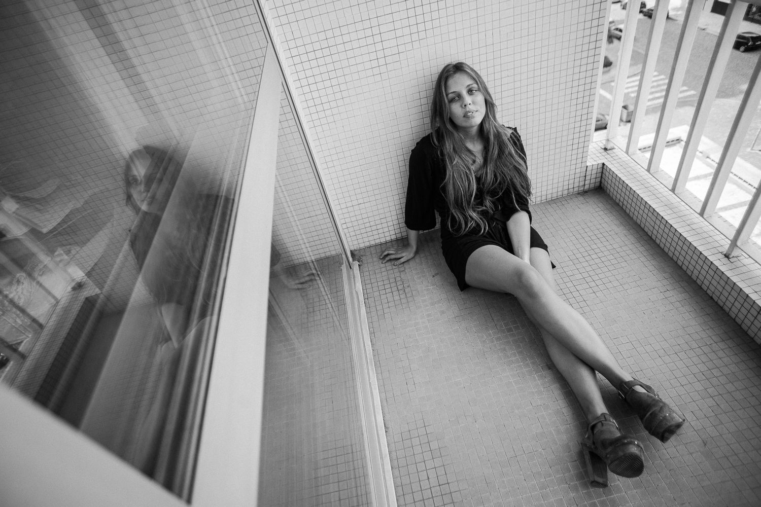 Person sits in corner of tiled room looking through window in dramatic black and white composition.