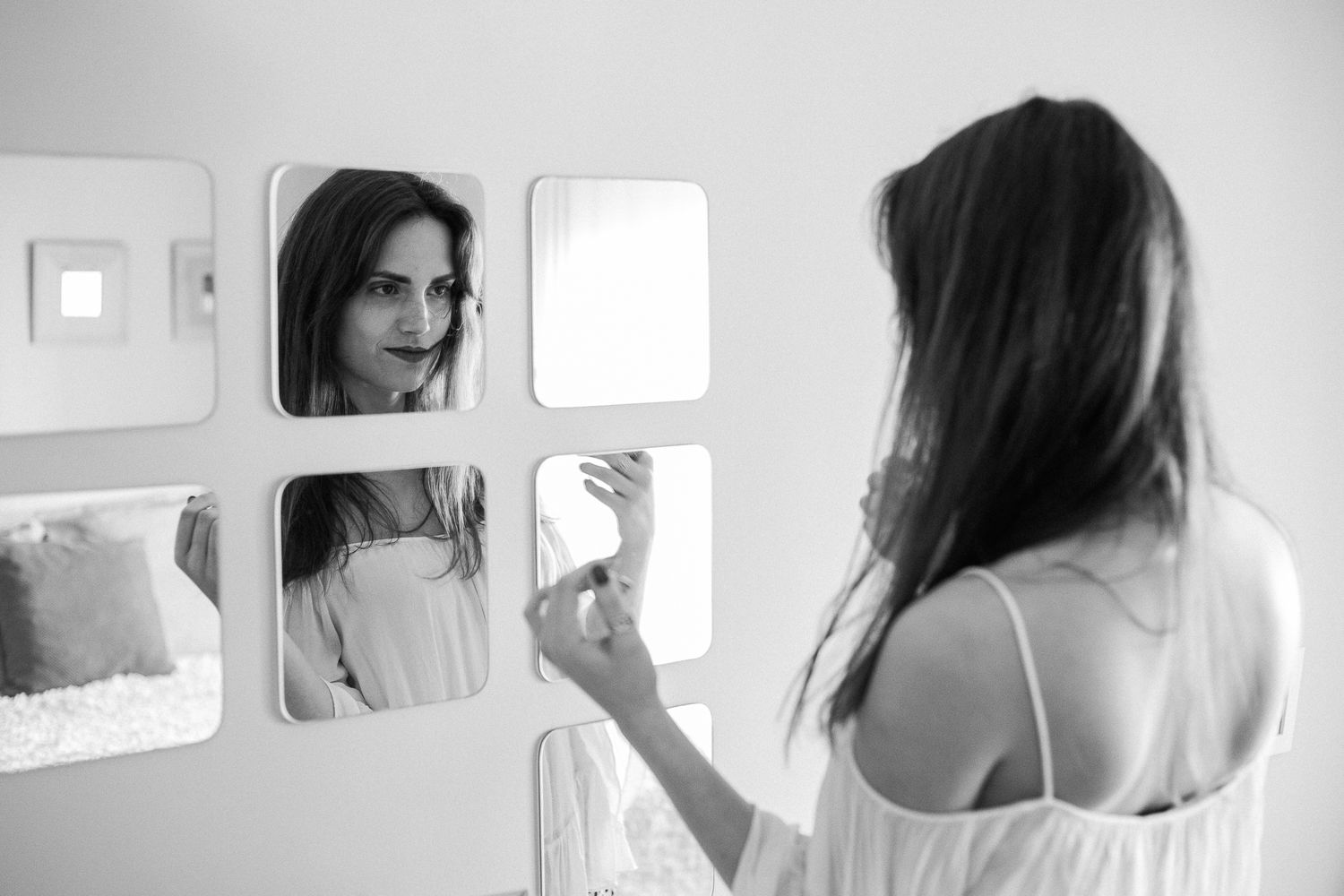 A person with long dark hair looks at their reflection in a multi-paneled bathroom mirror while wearing white clothing.