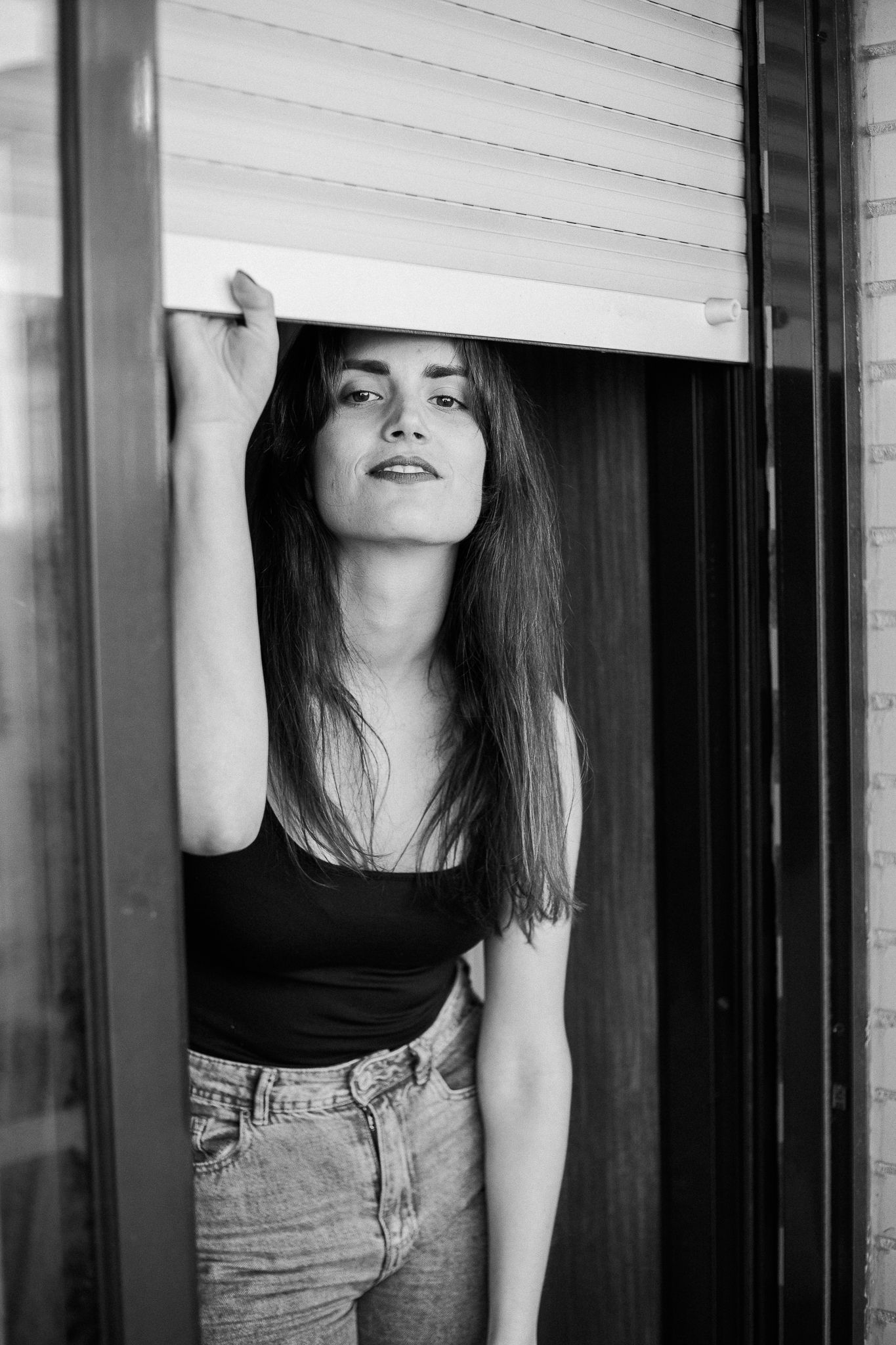 Black and white portrait of a person in a black tank top and jeans leaning against a window frame.