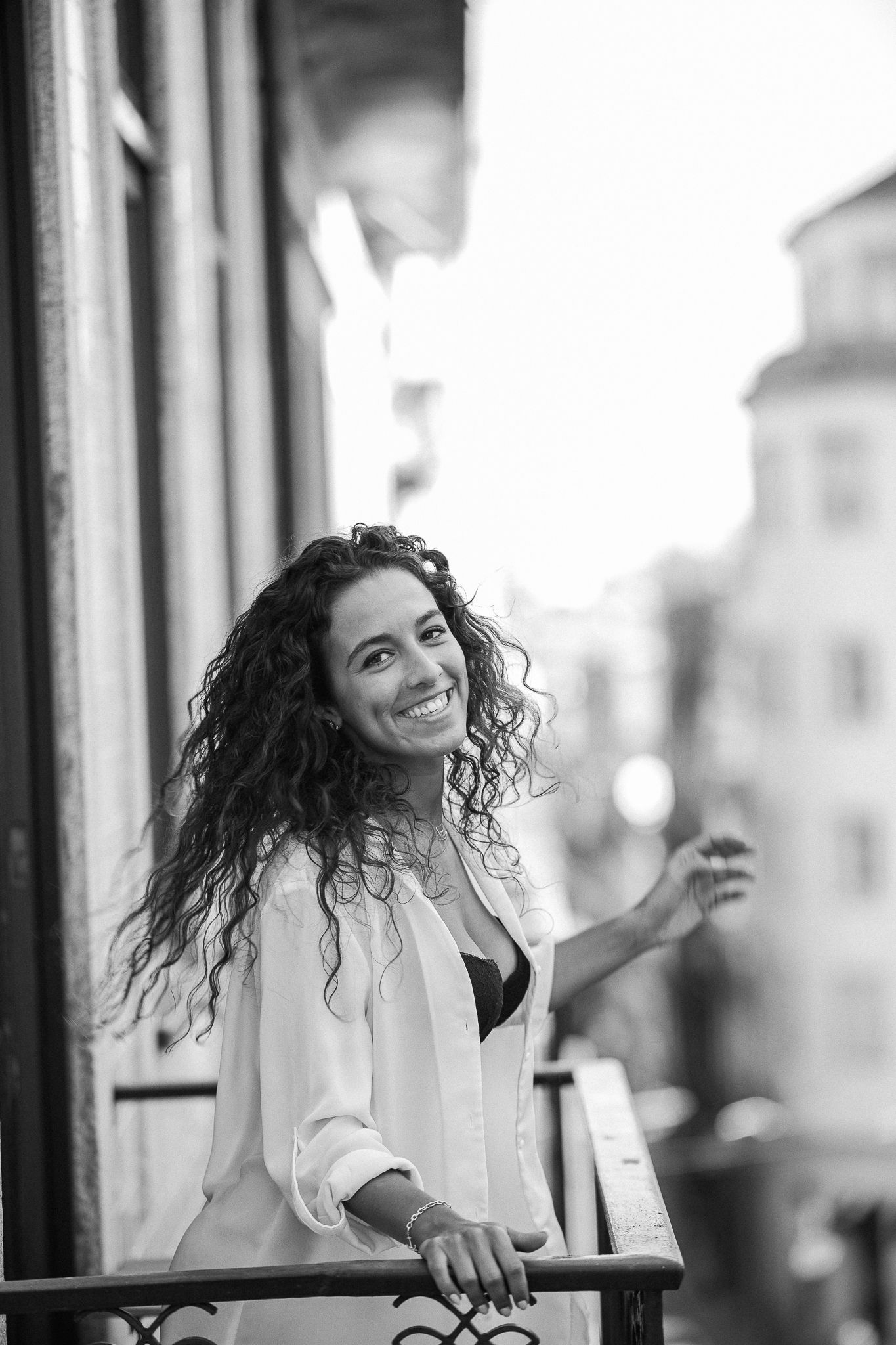 Black and white portrait of someone with curly hair smiling on a balcony wearing a light colored button-up shirt.