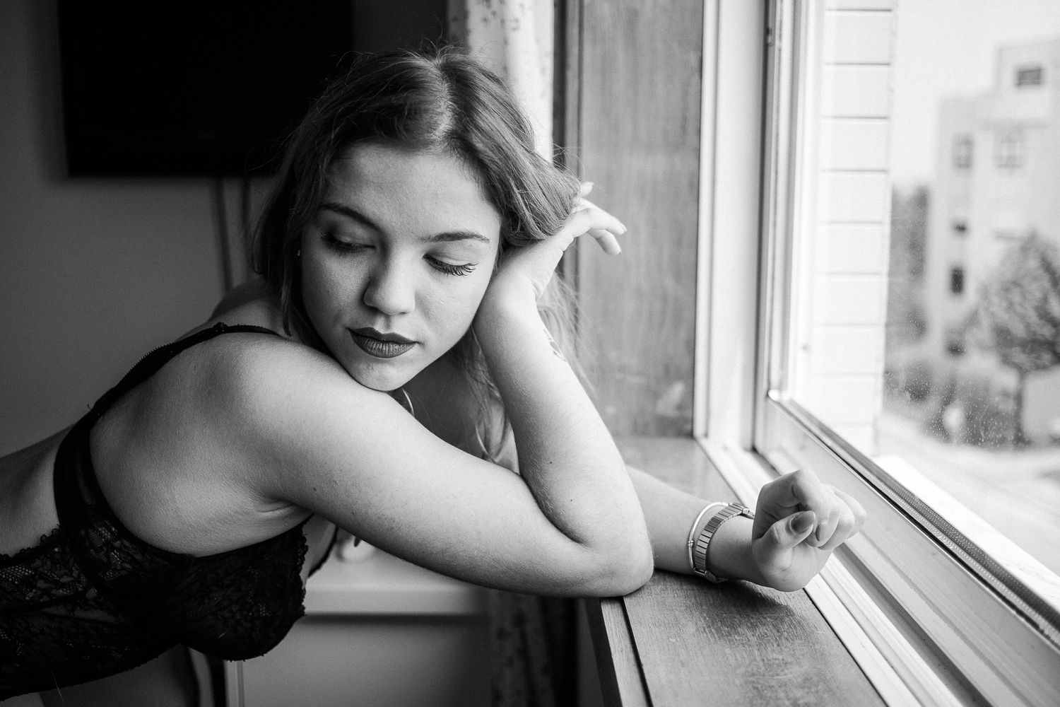 A black and white portrait of someone gazing thoughtfully out of a window while leaning on a windowsill.