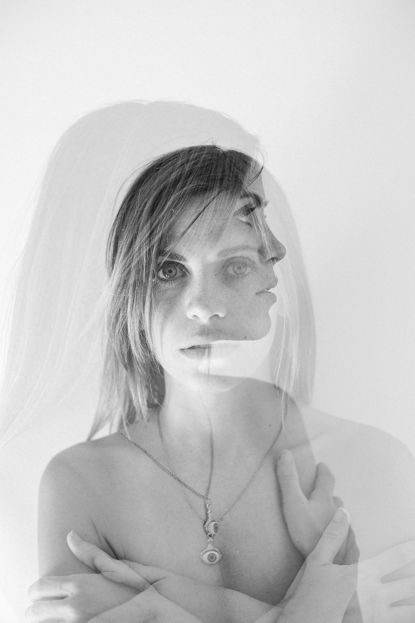 Artistic black and white portrait photograph showing ethereal double exposure effect with soft lighting and delicate necklace.