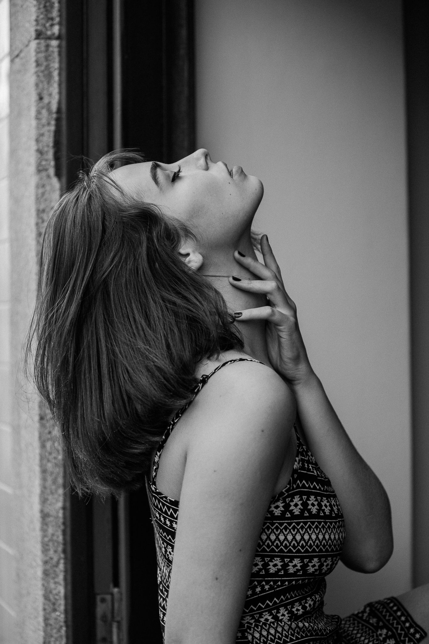 A black and white portrait of a person tilting their head back with eyes closed, wearing a patterned sleeveless top.