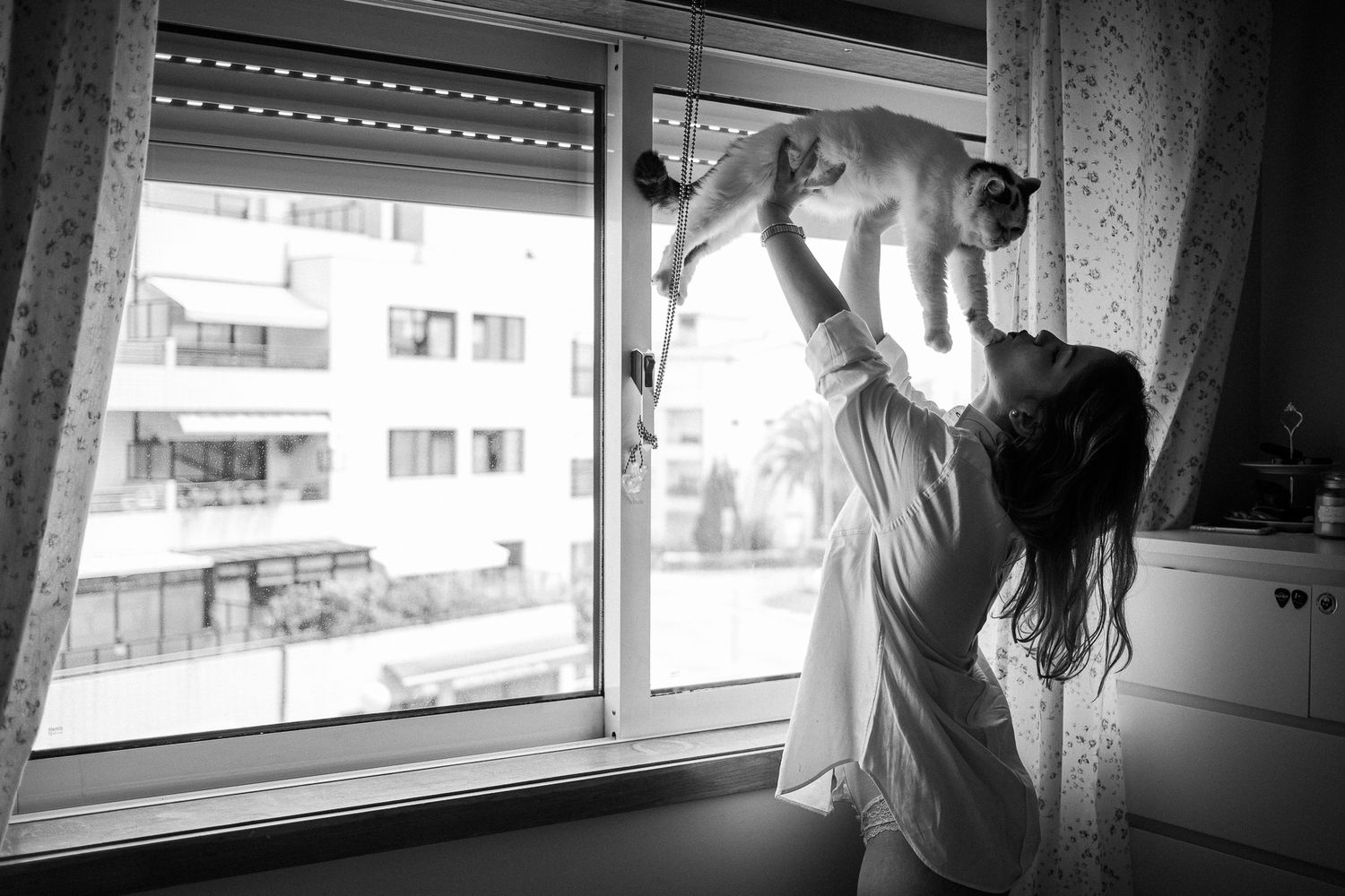 Black and white photo of a person looking through a window with reflective glass and urban buildings in background.