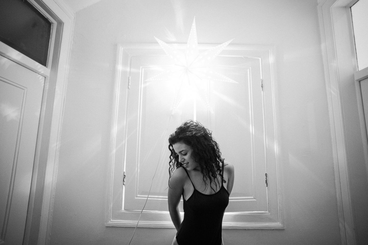 Black and white portrait of a person sitting against a white wall in moody lighting.
