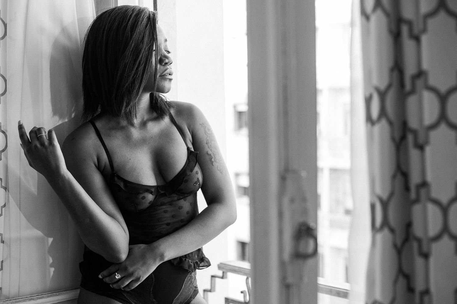 Black and white boudoir photo of woman in lingerie posing near window with natural lighting.