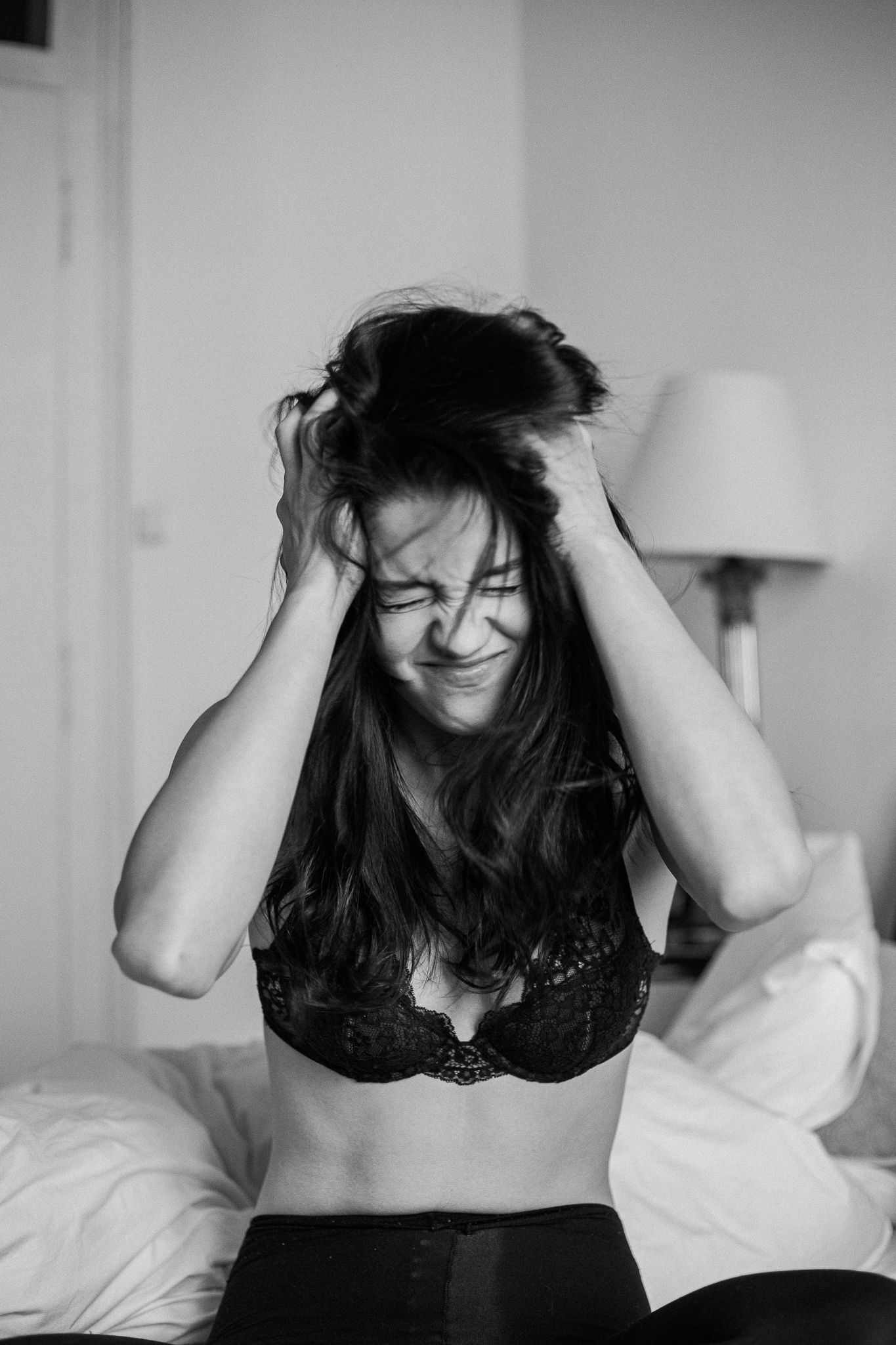 Black and white portrait of someone in a lace bralette running hands through messy hair while sitting on a bed.