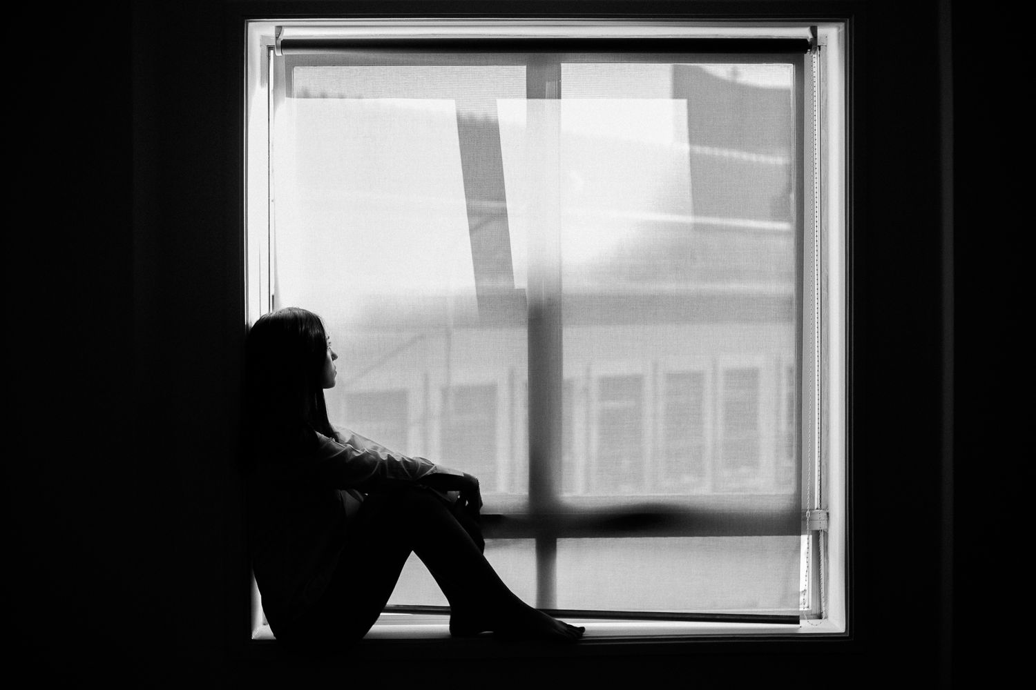 A silhouetted figure sits alone by a window in a dark room creating a moody black and white composition.