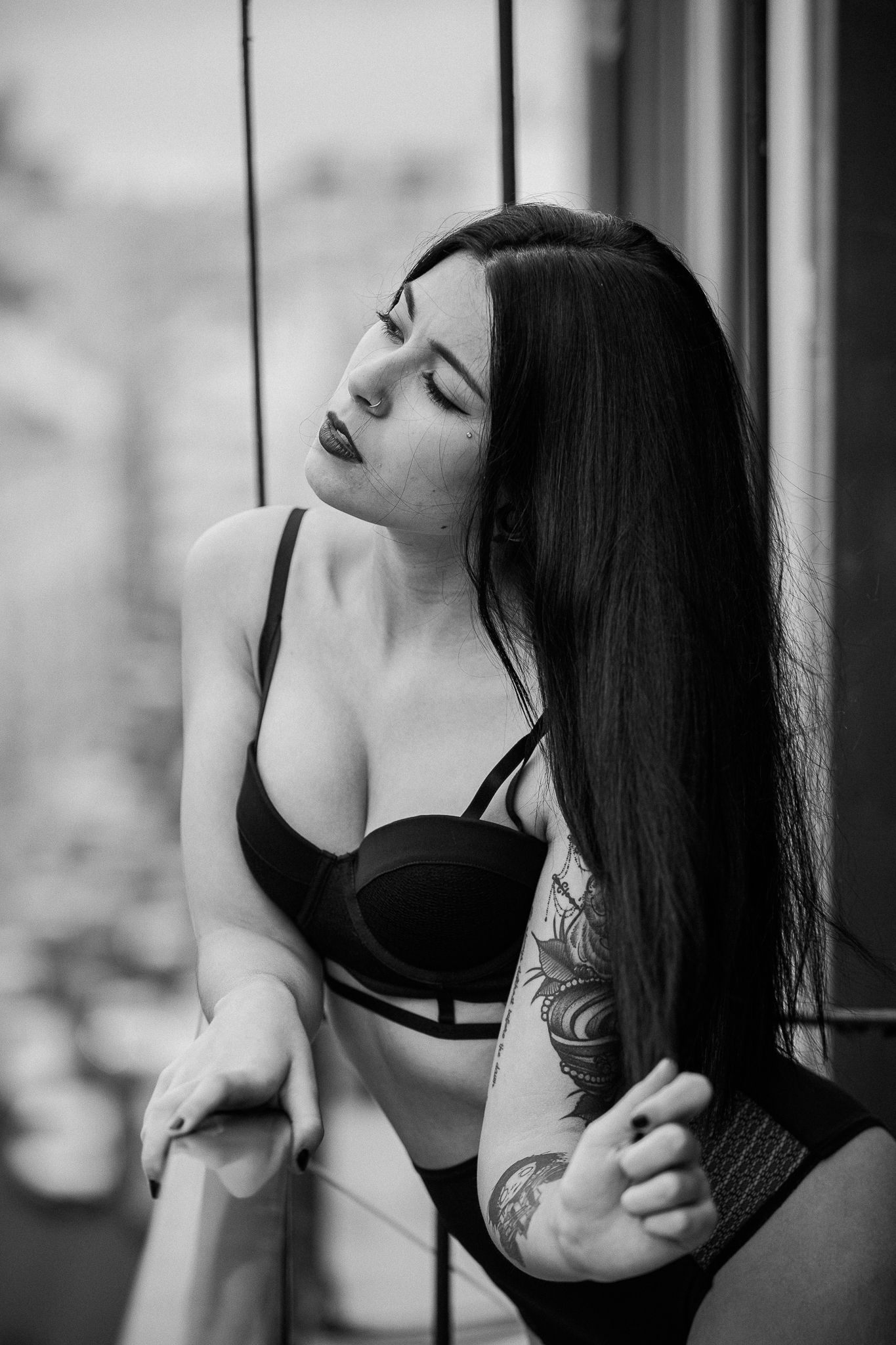 Artistic black and white boudoir photo of a woman with long dark hair and tattoos posing near a window.