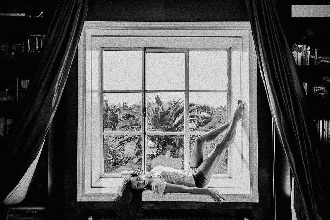 A person lounges on a windowsill in black and white, with dark curtains framing the window view.