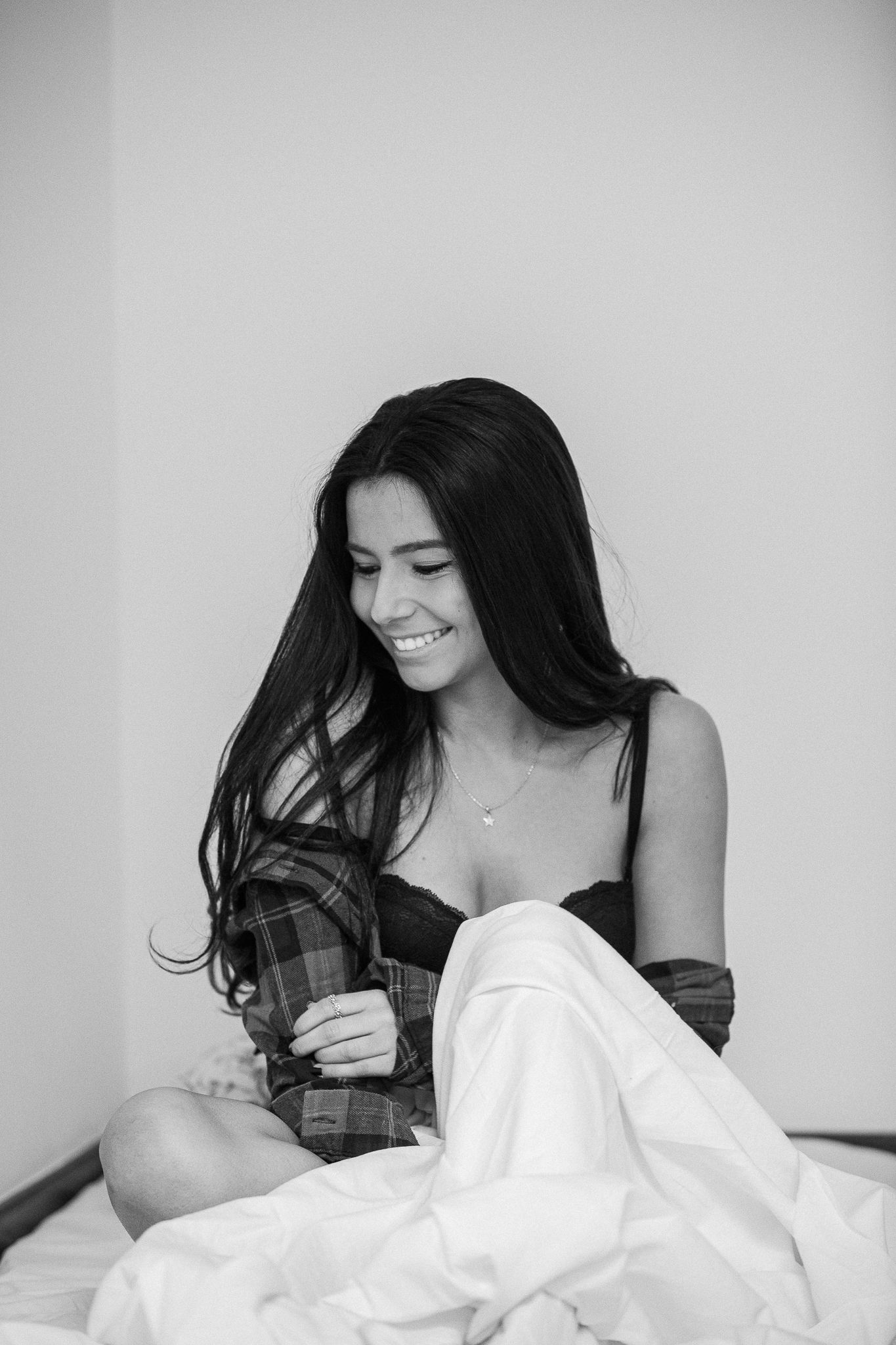 Black and white portrait of someone with long dark hair smiling while sitting on a bed with white sheets.