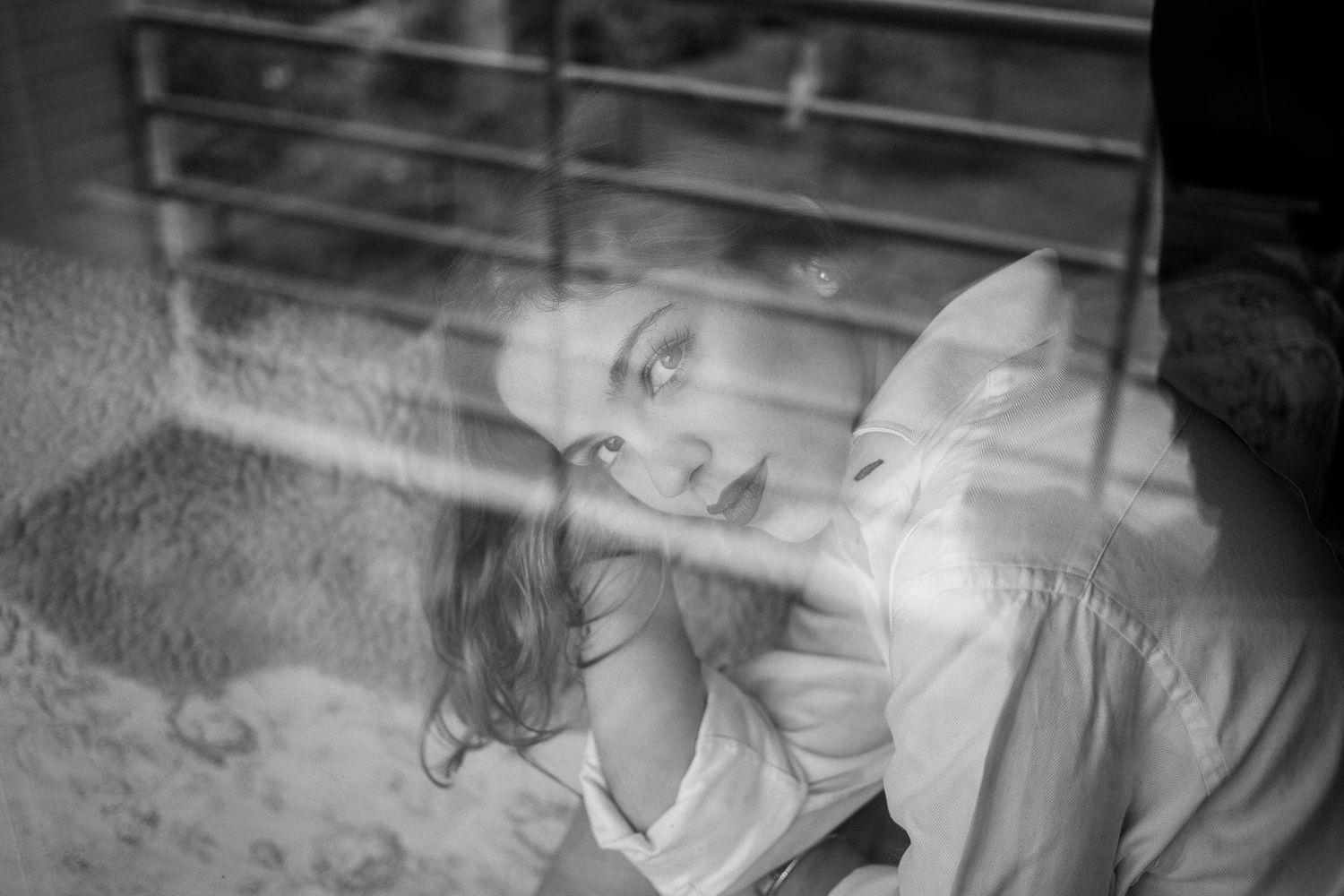 A dreamy black and white portrait through glass showing a resting figure in soft, ethereal lighting.