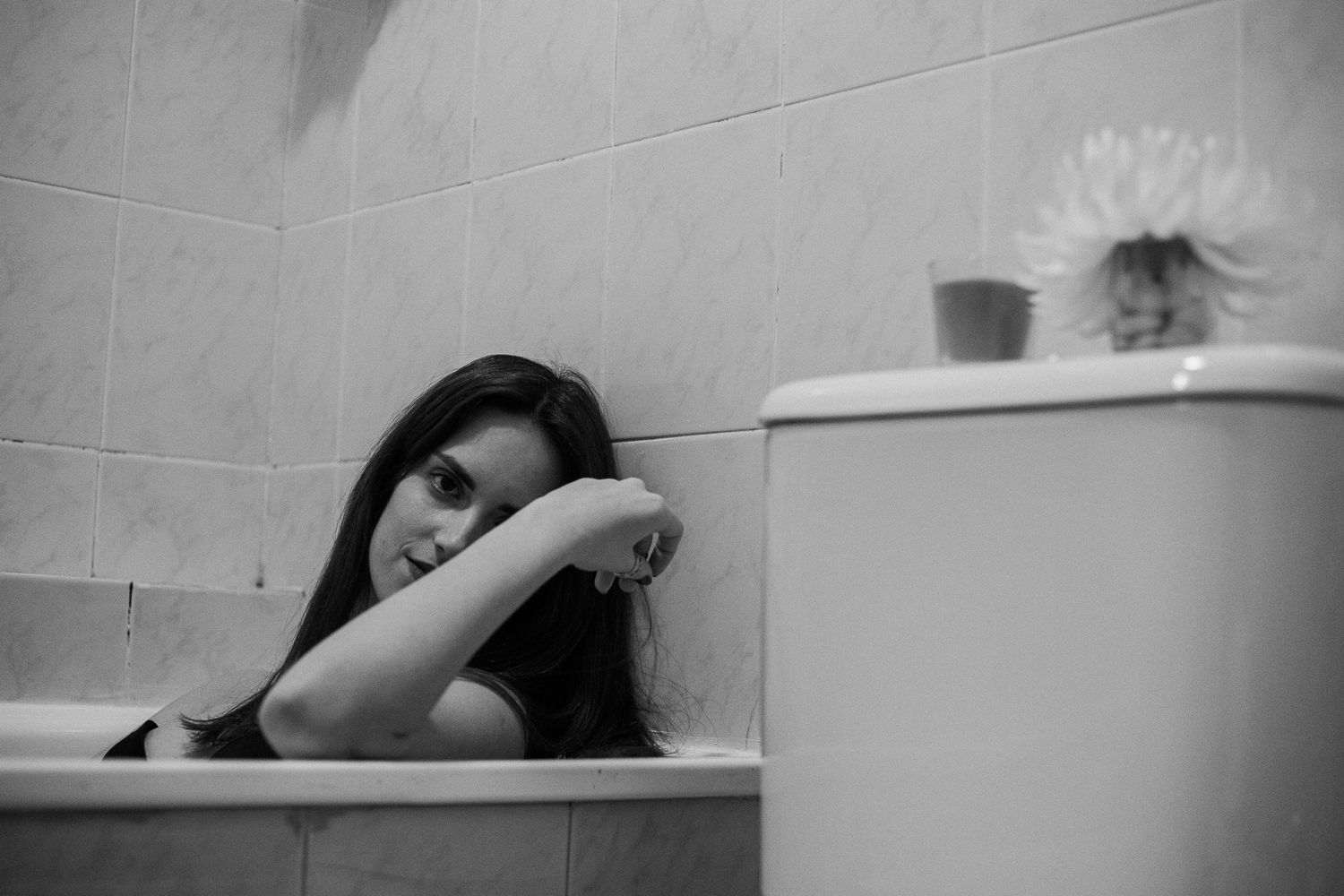 A black and white photo shows someone relaxing in a bathtub with a potted plant visible on the ledge.