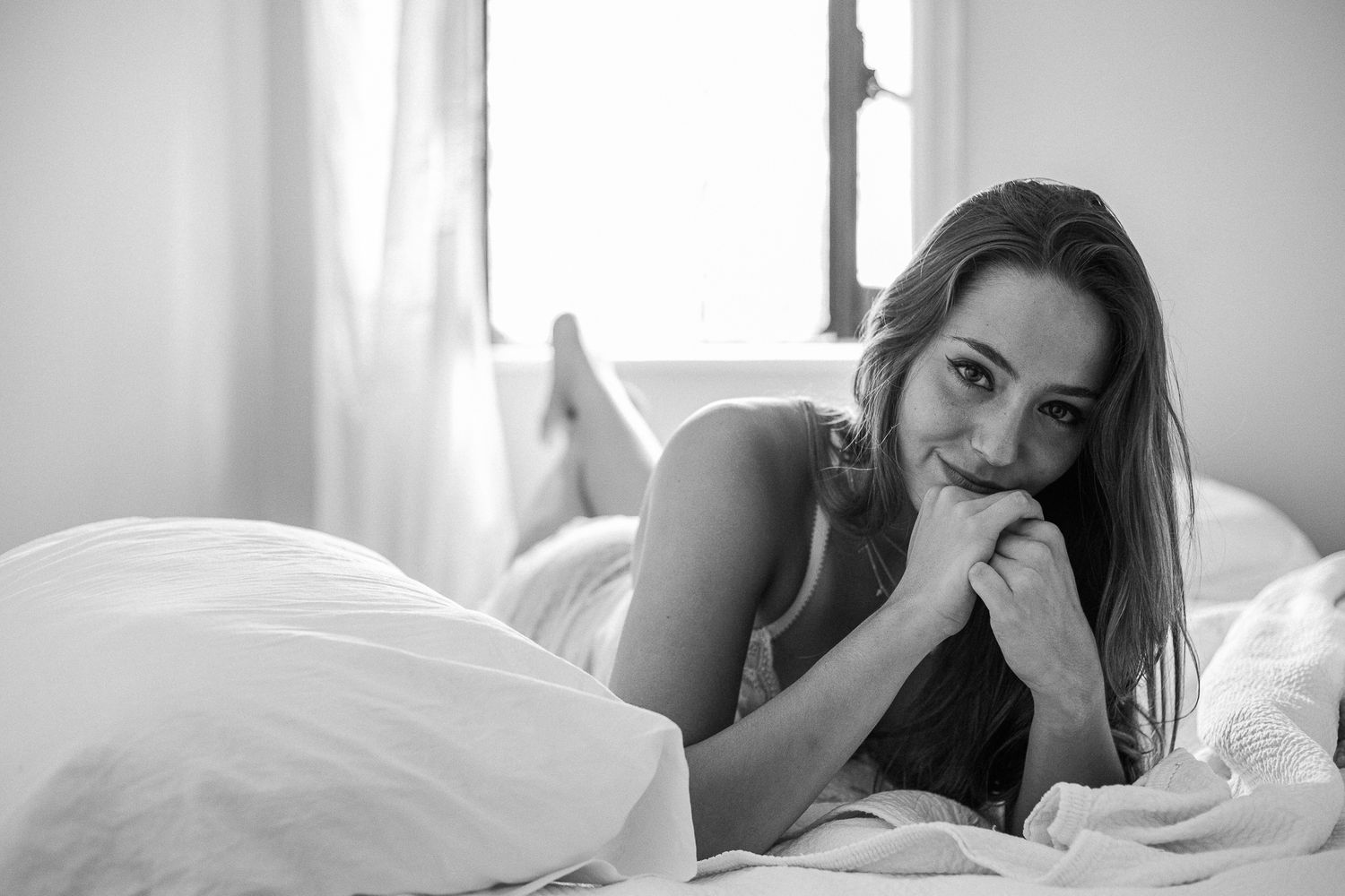 A person lounges seductively in white bedding near a bright window in a romantic boudoir setting.