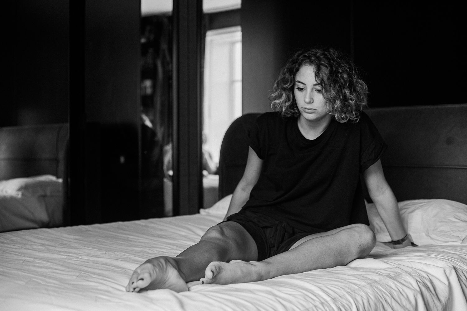 A person with curly hair wearing a black shirt sits casually on a bed in a black and white photograph.