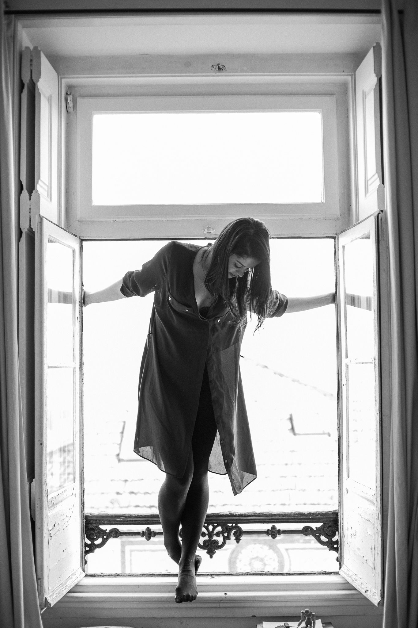 Black and white artistic photo of a figure balancing in a bright open window frame with ornate details below.