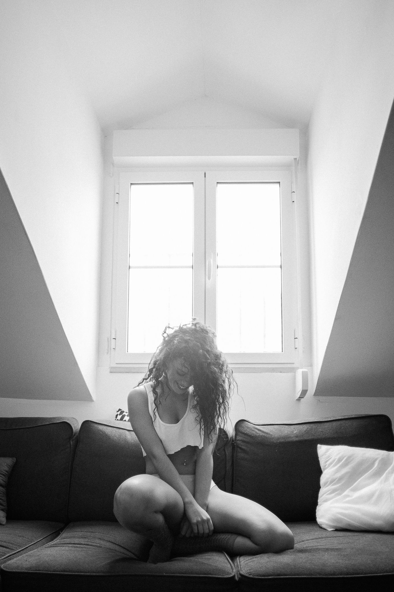 Black and white photo of a person sitting alone on a dark couch beneath a bright window in an attic room.