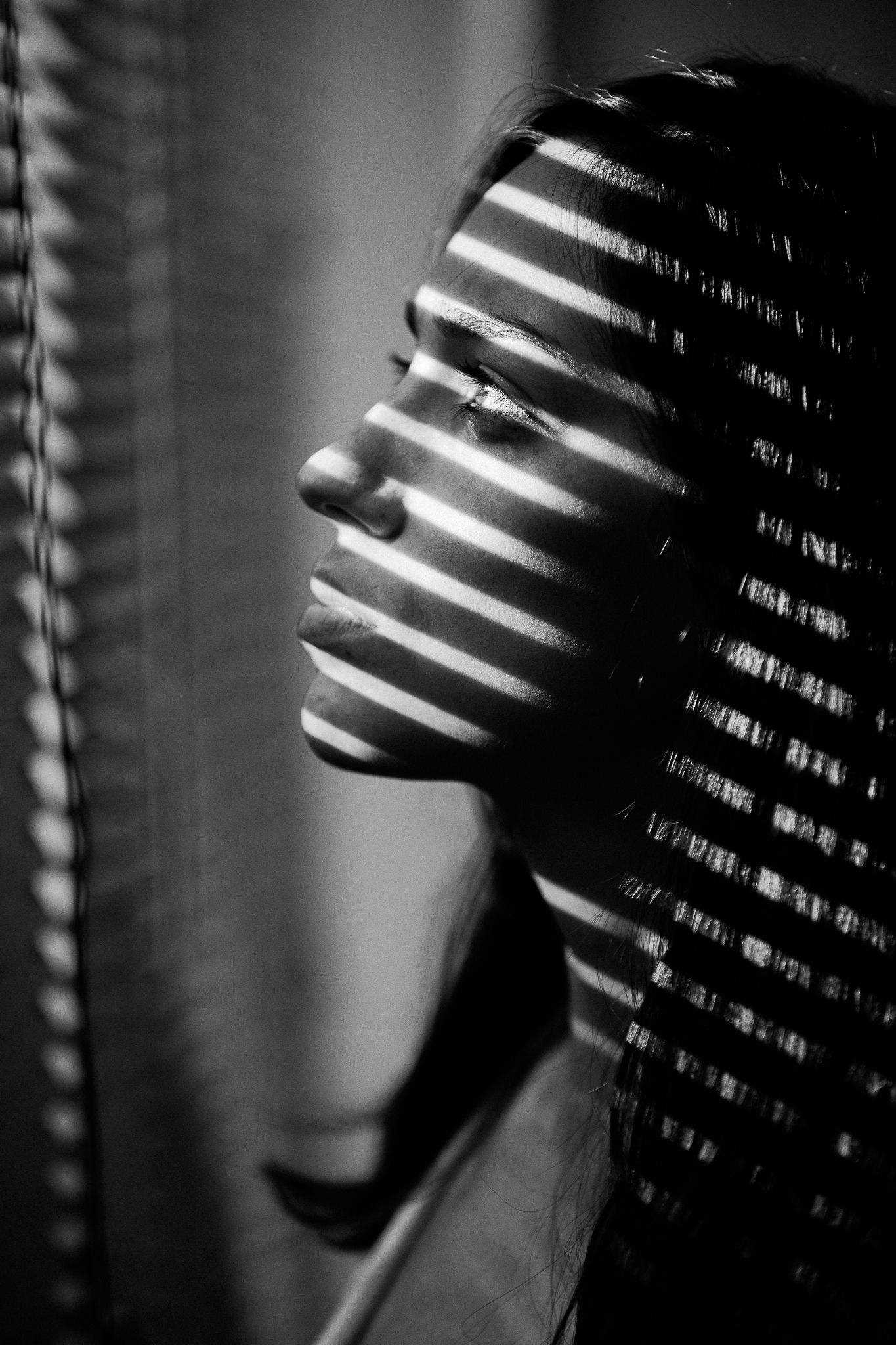 Black and white artistic portrait with dramatic shadows and venetian blind lighting effects creating a film noir atmosphere.