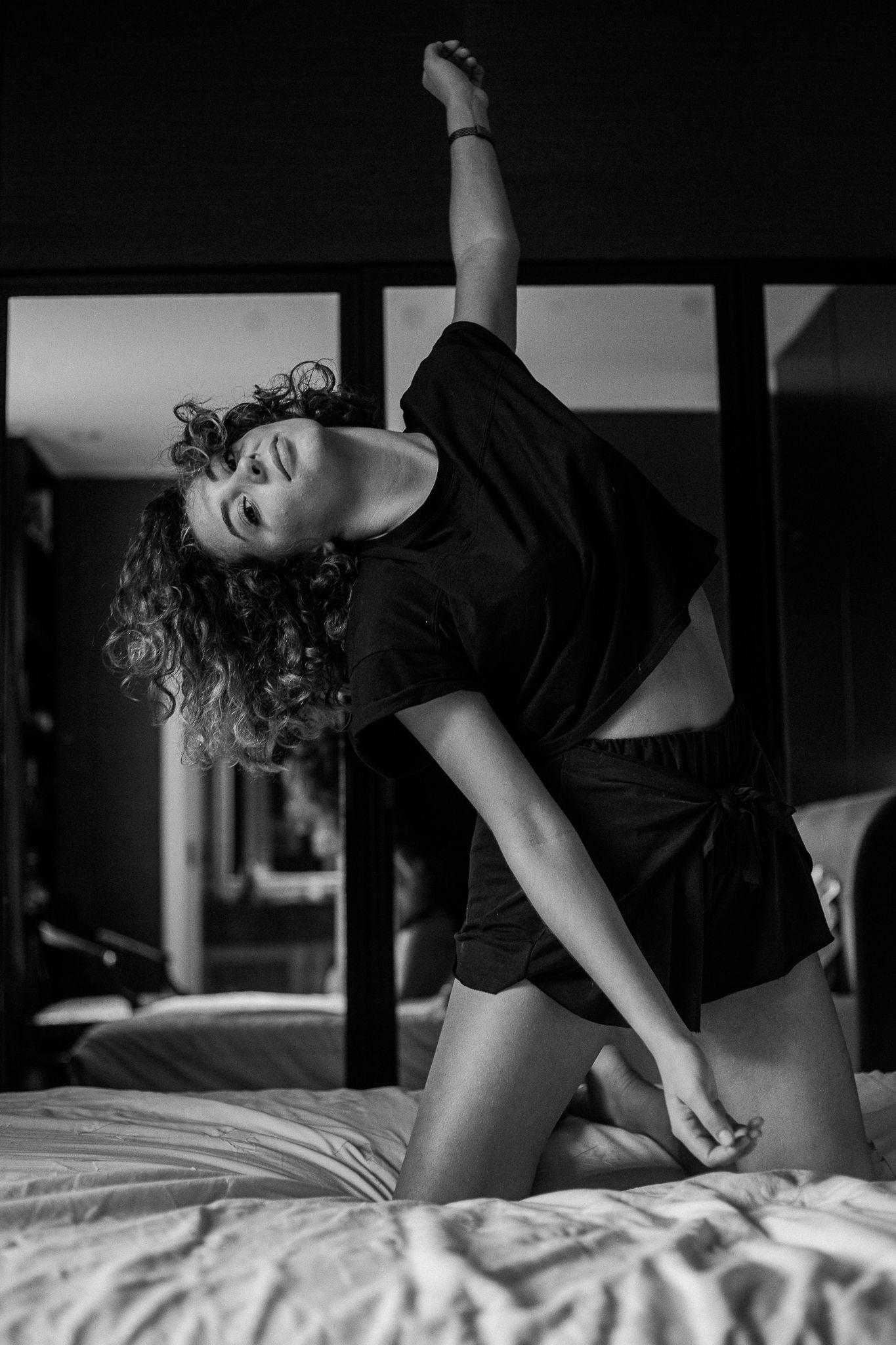 Black and white artistic portrait of a dancer in casual wear striking an expressive pose on a bed.