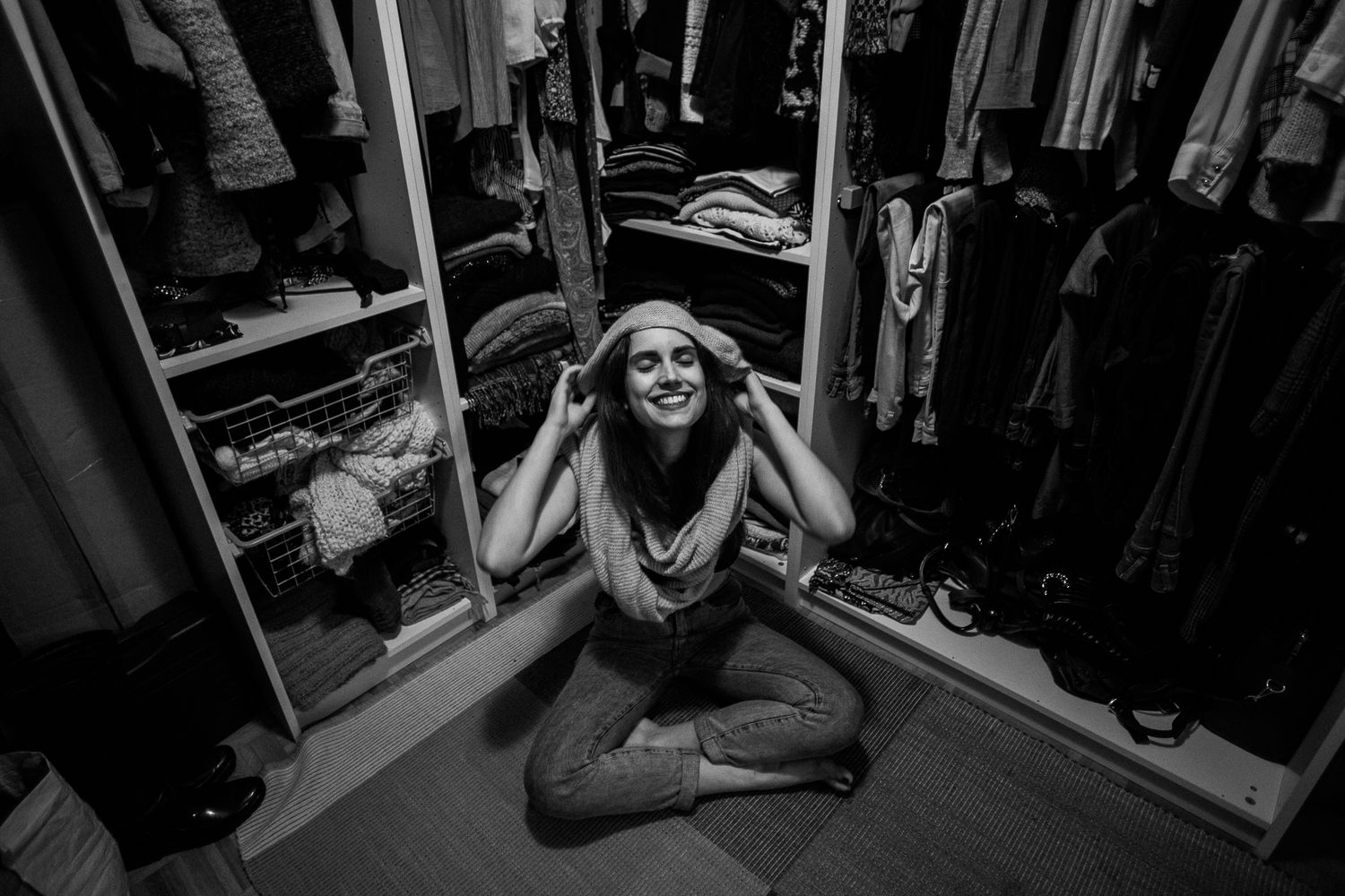Black and white photo of someone sitting cross-legged on floor in front of open closet with clothing.