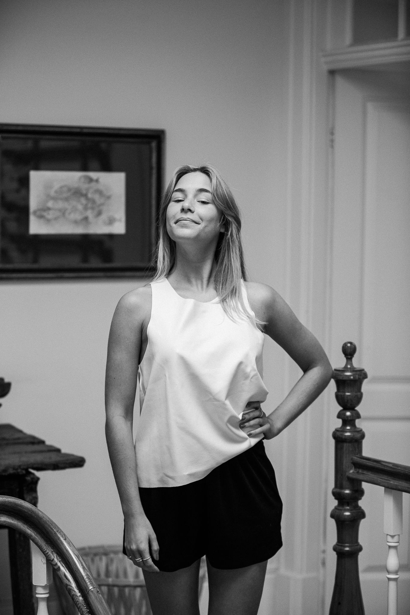 Black and white photo of someone in a white sleeveless top and dark shorts posing casually indoors with a relaxed smile.