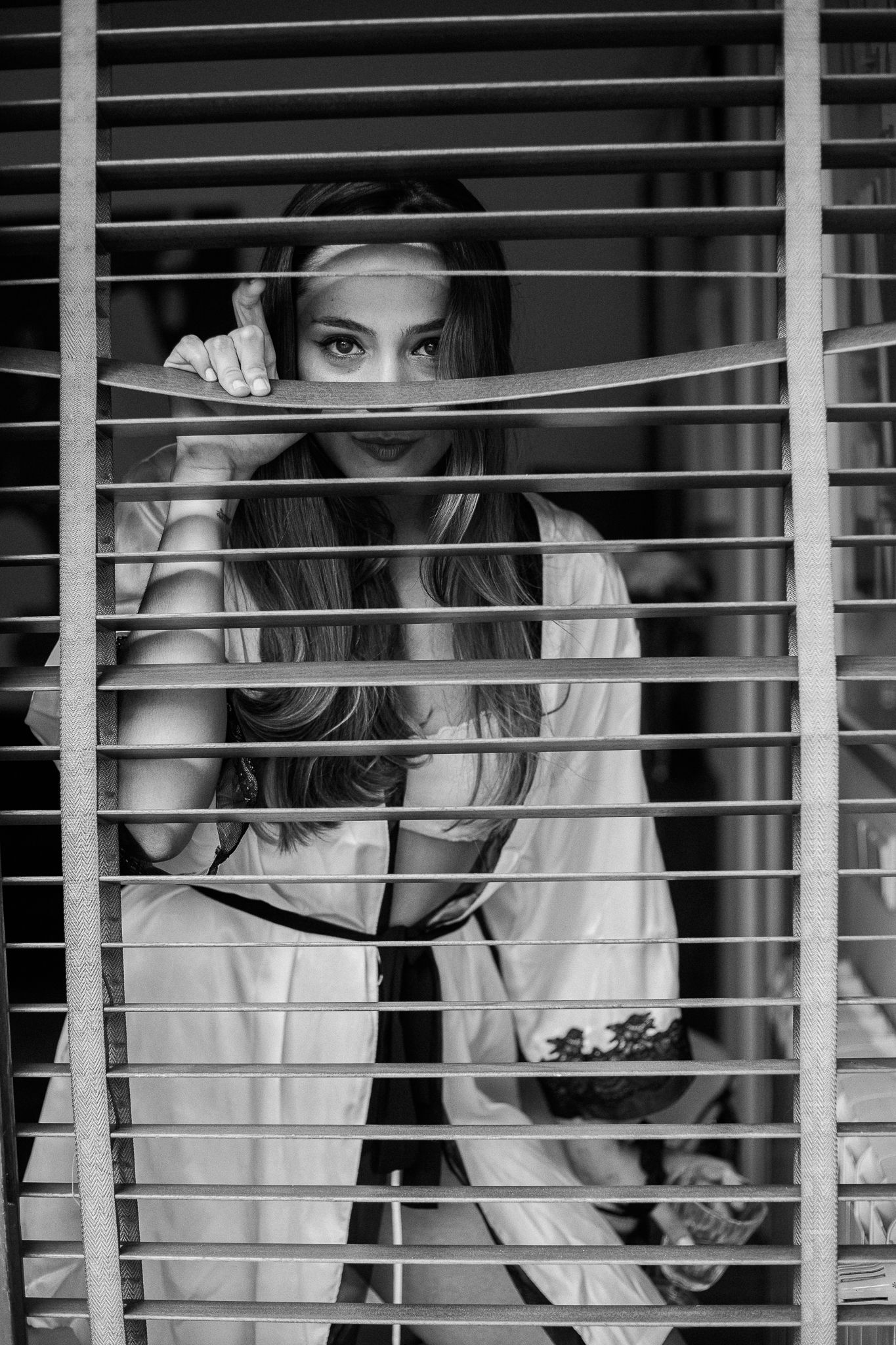 A black and white photo showing someone peeking through horizontal window blinds in a mysterious way.