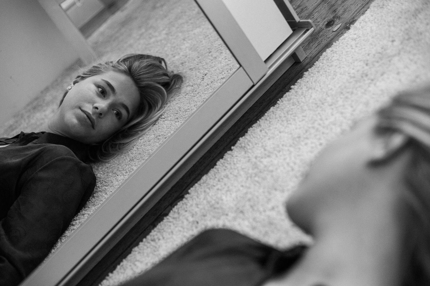 Black and white artistic reflection in mirror showing person lying on carpet with dramatic lighting and composition.