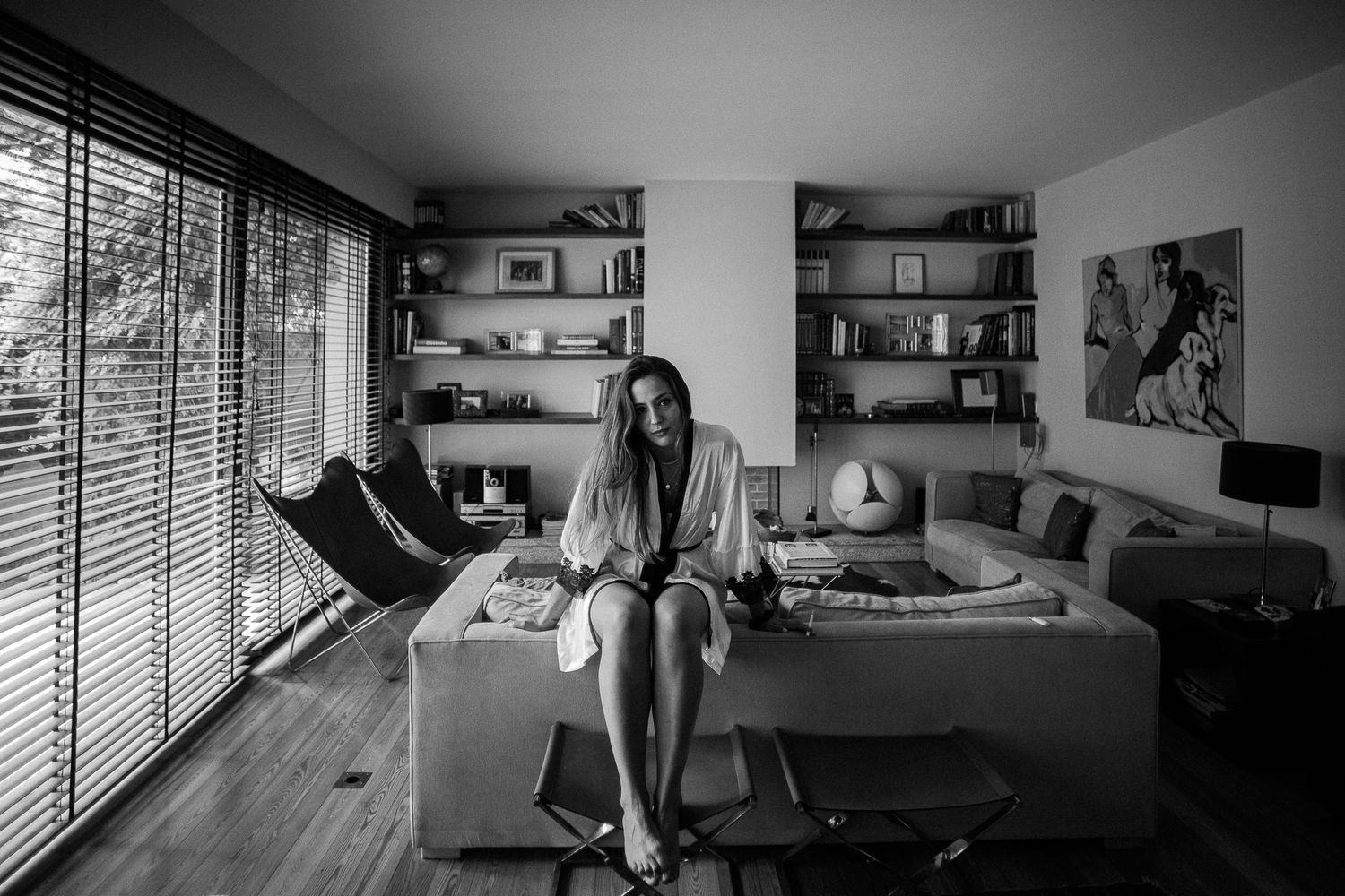 Black and white photo of a person sitting on a couch in a modern living room with bookshelves and large windows.