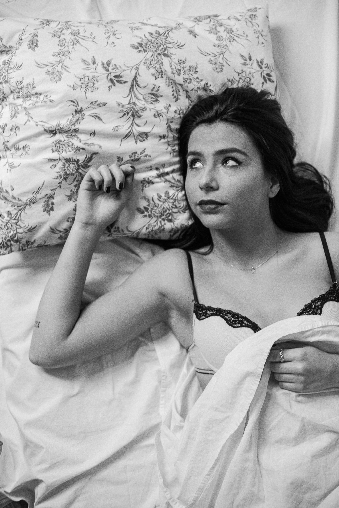 Black and white boudoir photo showing someone reclining on floral bedding while gazing upward in a dreamy pose.