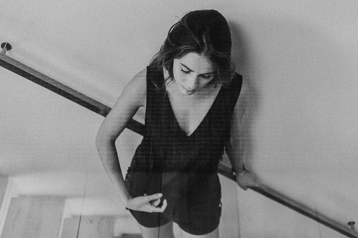 A person in a black dress looks down at their phone while standing against a white wall in a black and white photo.