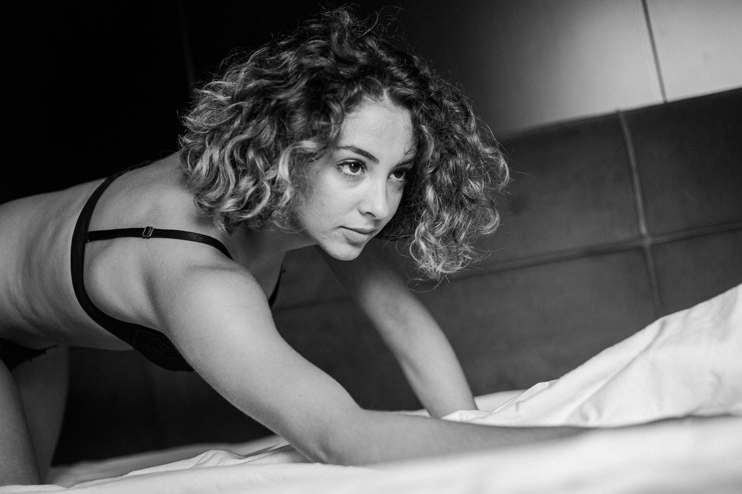 Black and white boudoir portrait of a model with curly hair posing on a bed.