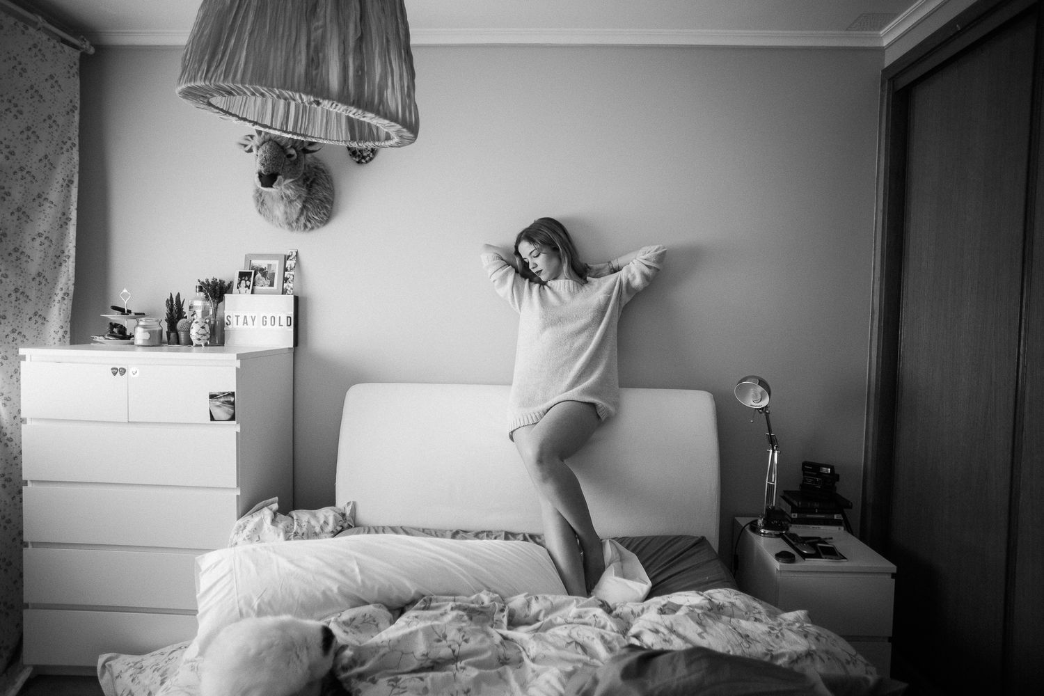 A person lies in bed with messy white sheets in a dimly lit bedroom interior.