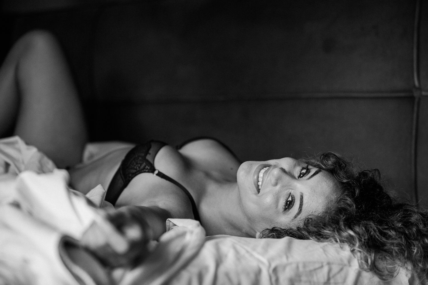 Black and white boudoir photograph showing a person reclining on white bedding in intimate apparel.