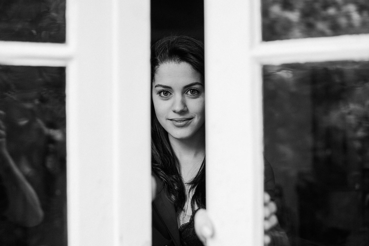 A portrait photo through white wooden fence slats shows someone looking at the camera with a gentle smile.