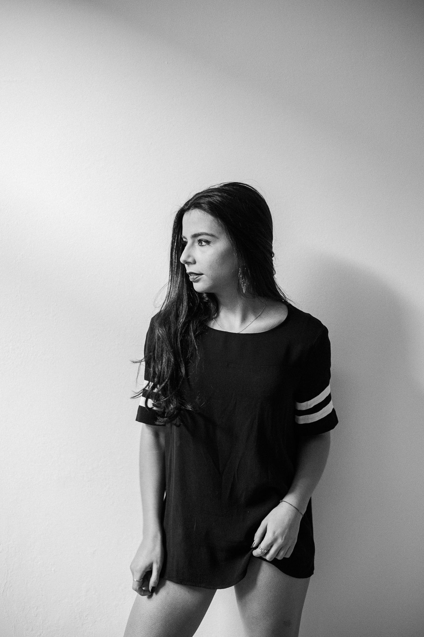 Black and white portrait of someone in casual dark top with striped sleeves looking to the side against plain wall.