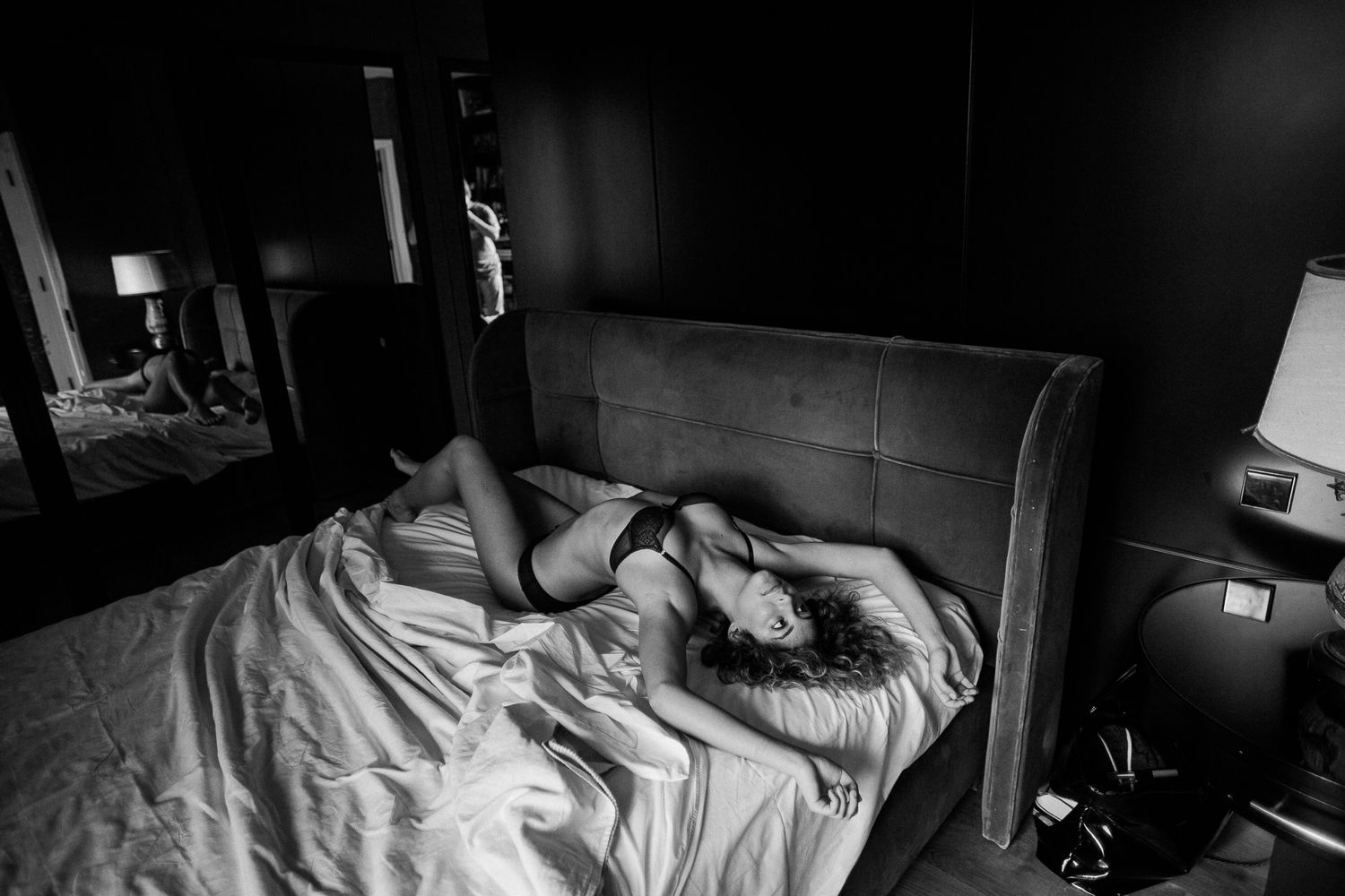 Black and white photograph showing a messy unmade bed with rumpled sheets and pillows in dim lighting.