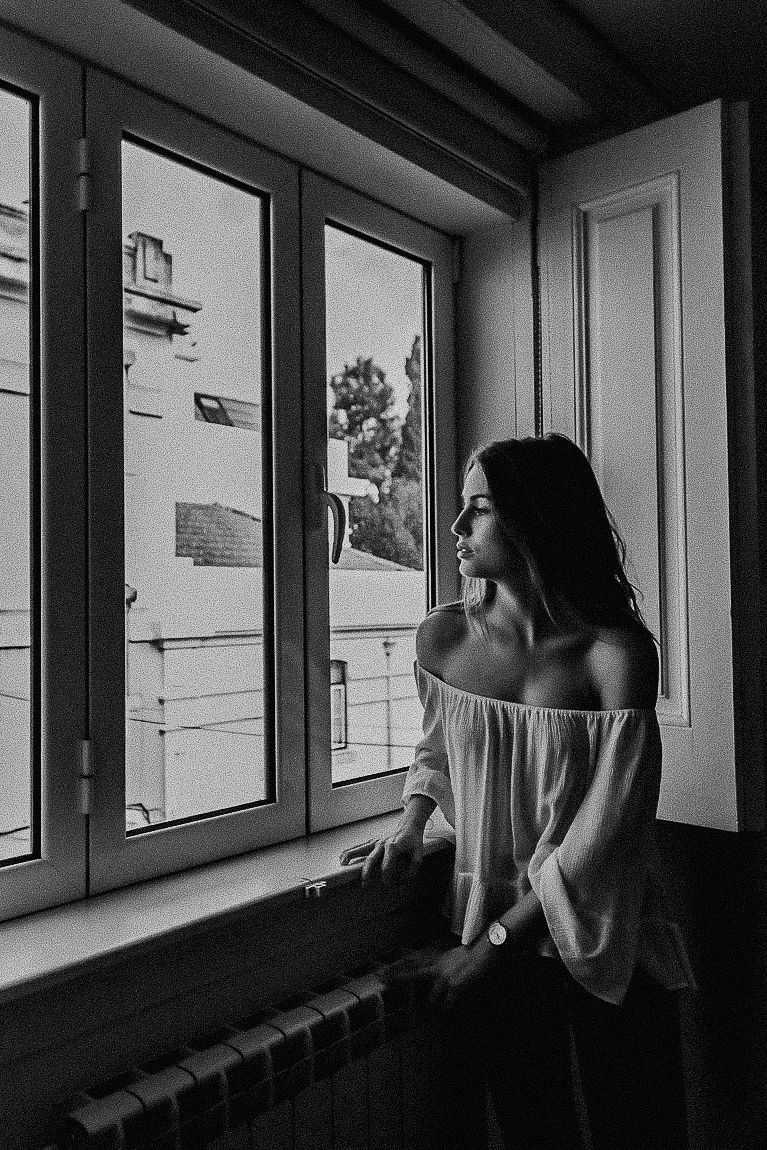 A black and white photograph shows a figure gazing out of a row of windows in a minimalist interior space.
