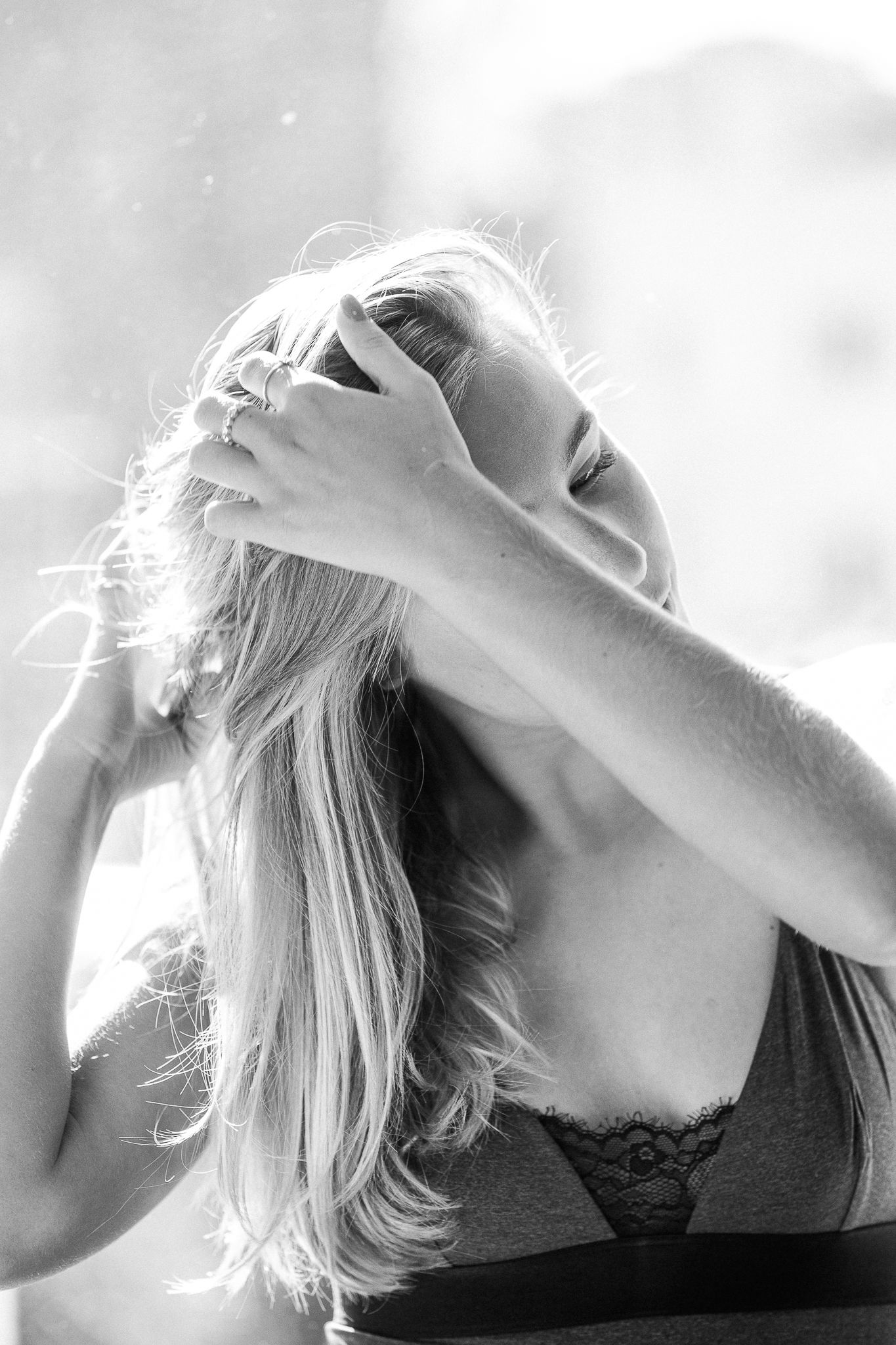 Black and white artistic portrait of someone with long blonde hair in lingerie throwing their head back with sunlit glow.