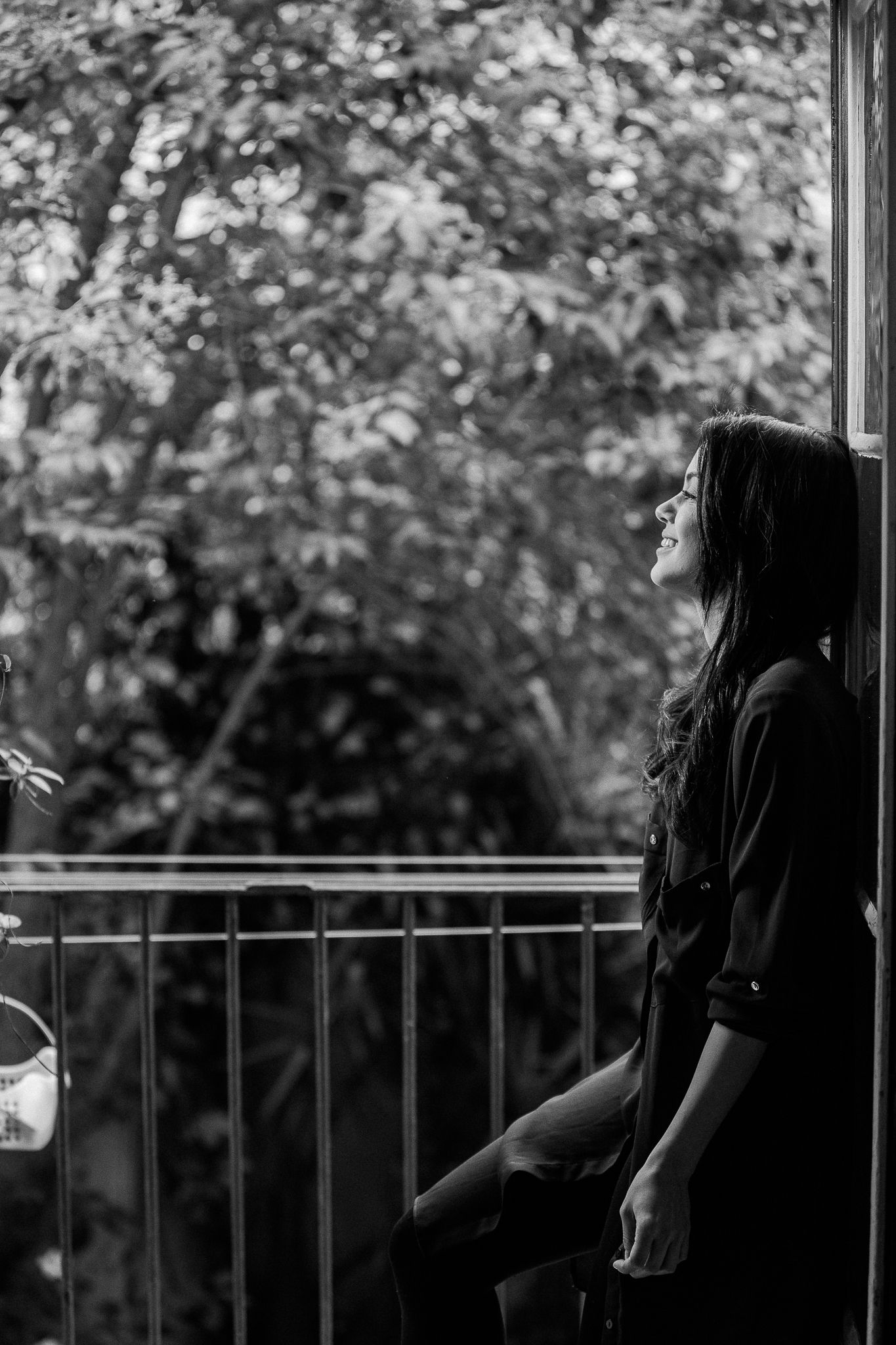 A person in dark clothing stands thoughtfully on a balcony against a backdrop of trees in black and white.