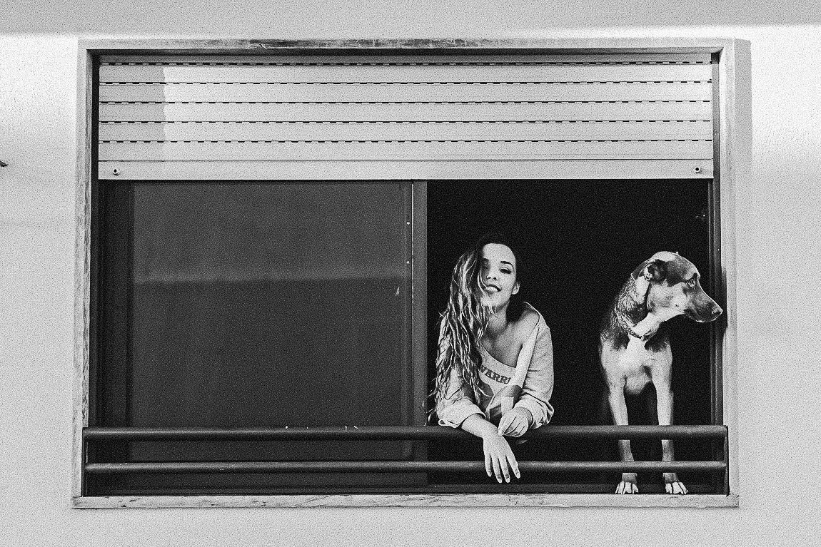 Black and white photograph showing people looking out of apartment windows on a city street.