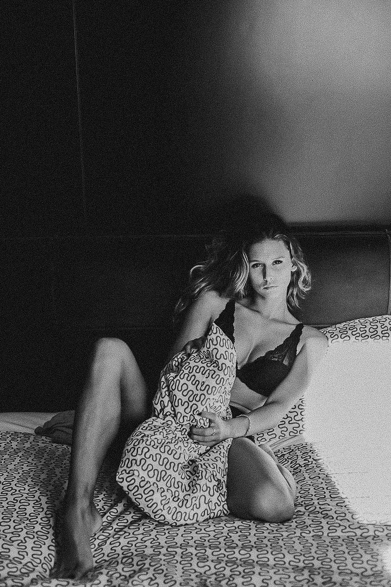 Black and white boudoir photograph showing a person in a patterned dress reclining on a textured bedspread.