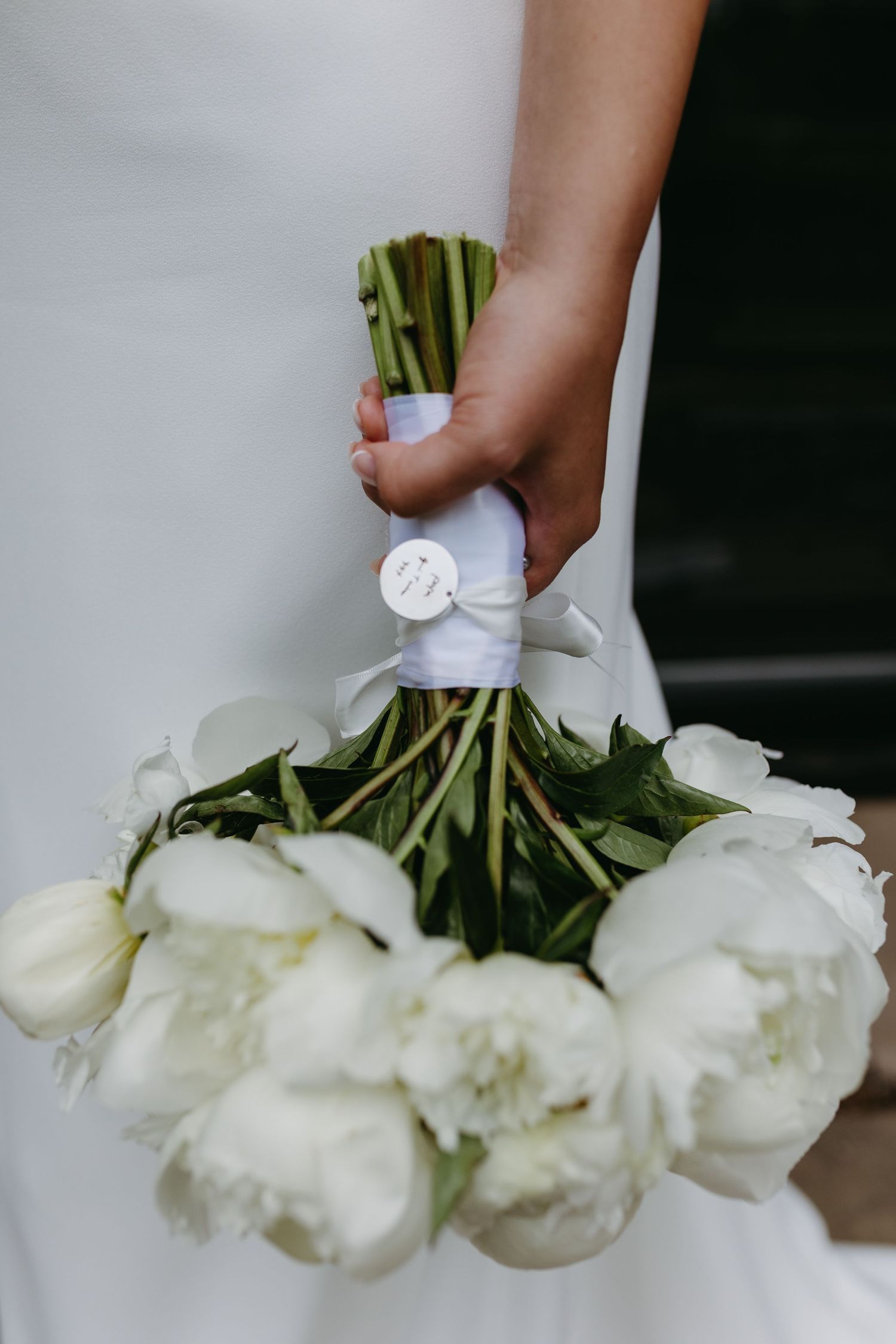 A hand holding an elegant bouquet of white peonies wrapped with a white silk ribbon.