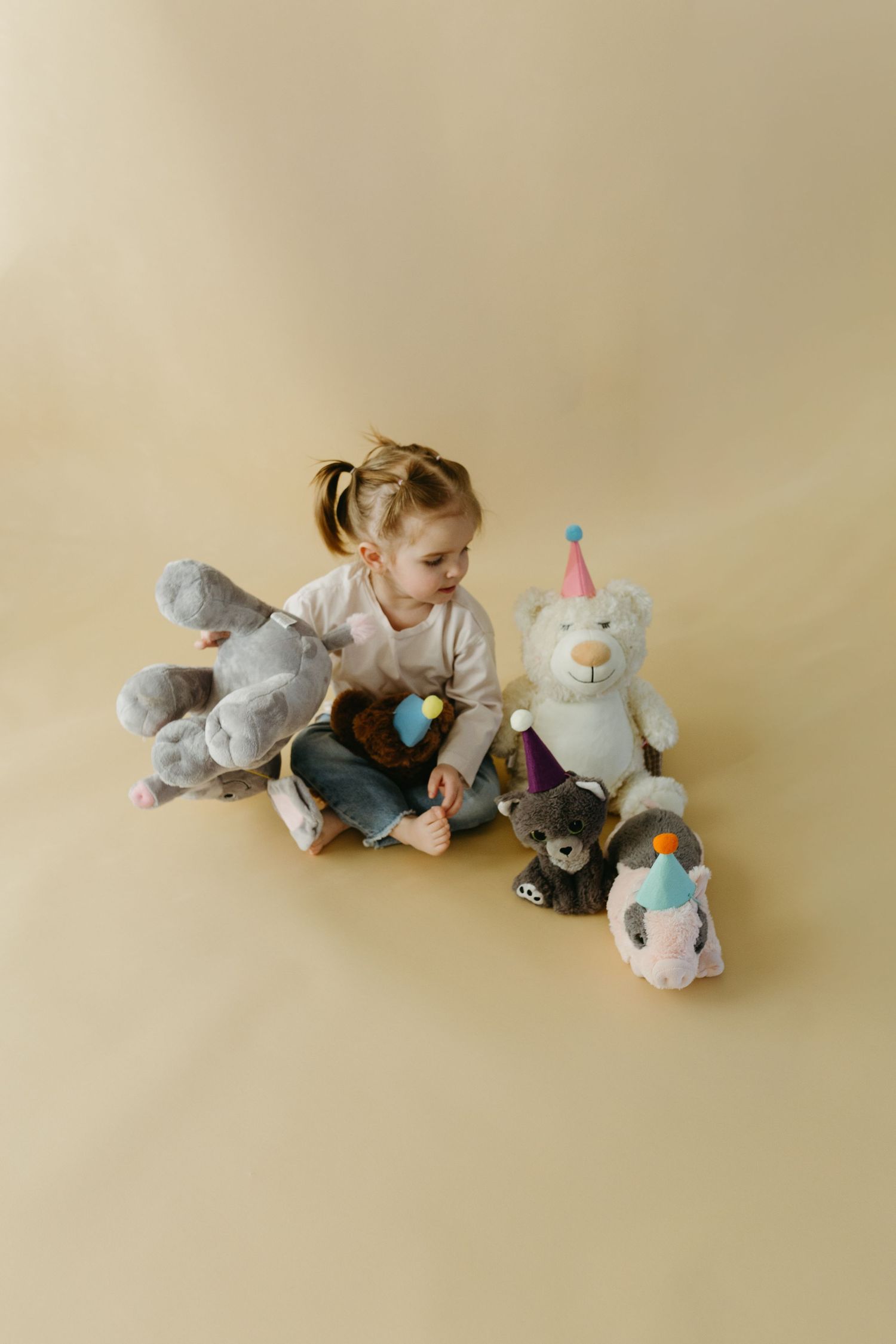 A small child sits surrounded by plush stuffed animals including bears and unicorns on a beige background.
