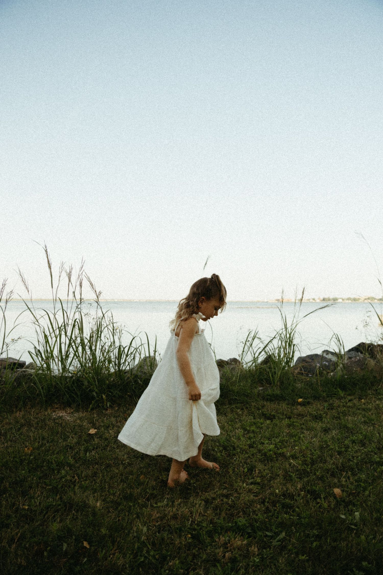 A person in a white flowing dress walks through tall grass near the ocean at sunset creating a dreamy silhouette.